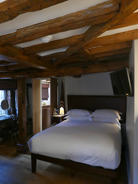 A room in an old cottage, with exposed wooden beams across the ceiling. The bed has a white cover.