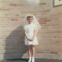 An image of the author in her first communion dress in front of the exterior wall of the church.