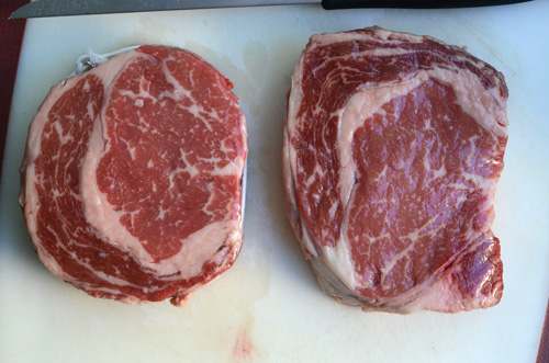 On the left, a competition style trimmed ribeye and on the right, an untrimmed ribeye steak.