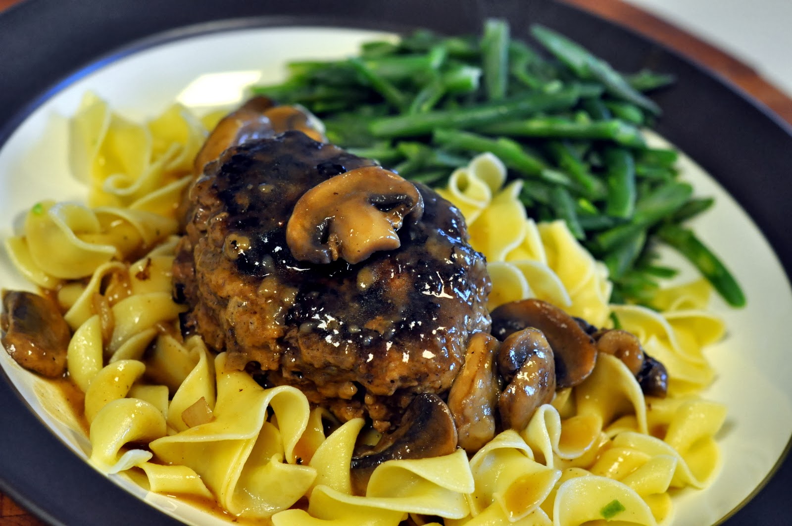 Salisbury steak with mushrooms over wide egg noodles and served with green beans. Food is on a blue and white plate.