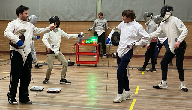 Four pistes of fencers fencing epee and sabre in the wooden floored fencing hall! Hmm, where are the foilists? They must be out of shot. Anyway, some fencers are on wired and some on wireless scoring gear. On the piste closest to the back wall the Favero scoring box registers a touch for fencer right, lighting up a green light. On the piste closest to us, two smiling fencers with their masks off get ready to plug in their small individual relay boxes before beginning their bout.