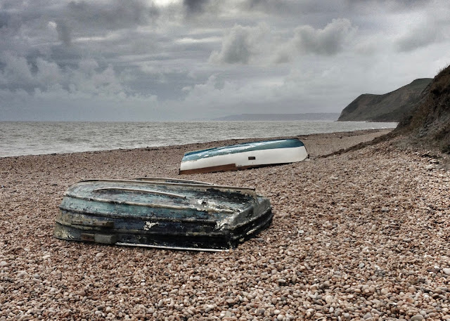 A pebble beach with grey skies and 2 rowing boats upside down on the pebbles