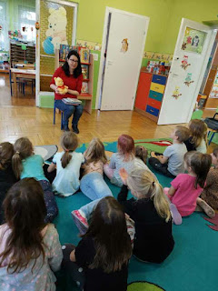Pani bibliotekarka trzyma w ręku maskotkę Kubusia Puchatka, dzieci siedzą na dywanie. Tło: sala przedszkolna.
