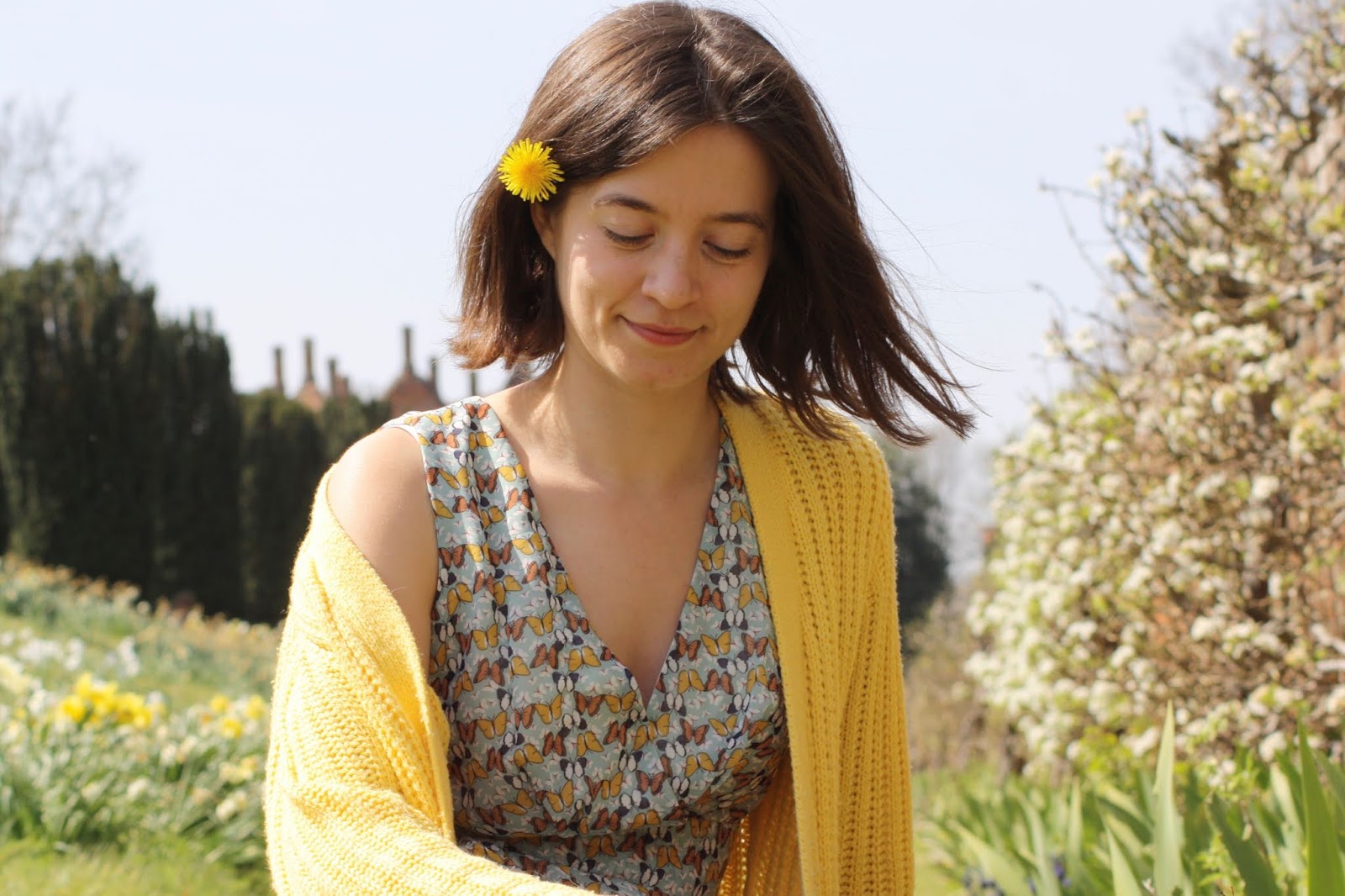 Abbey, wearing a butterfly print dress and yellow cardigan, smiles as she sits facing the camera with a dandelion tucked in her brown hair