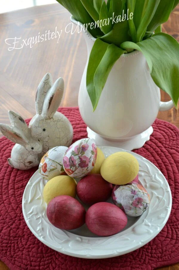 Decoupaged and painted Easter eggs on table with vase and bunny figures.