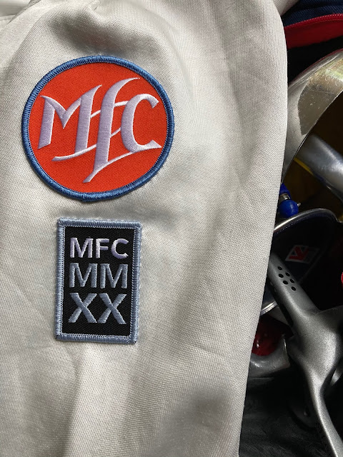 Close up photo of white fencing jacket sleeve with embroidered patches sewn to it: a club patch and 2020 commemorative patch. The Club patch is an orange circle with purple edge and the MFC logo in white. The 2020 patch is below it, a smaller vertical rectangle, black with MFC in white and MM and then XX below that in pewter. The patch has a pewter edge.