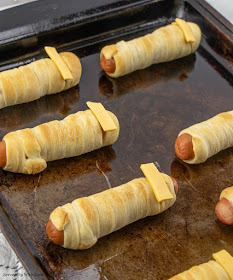 Hot dog wrapped in crescent roll strips to make a mummy lined up on baking sheet with cheese