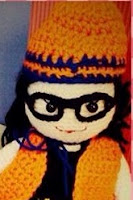 Close up of Kwokkie Doll's head and shoulders, wearing an orange beanie with blue trim, orange vest and black top. The blue strands around her neck belong to a cape she is also wearing.