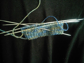 The cuff of a ribbed sock live on double-pointed needles.   Yarn is a blue flecked with deep gold. 