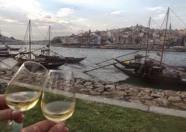 Cuas mãos brindando com copos de viho do Porto nas margens do rio Douro, barcos e a cidade do Porto