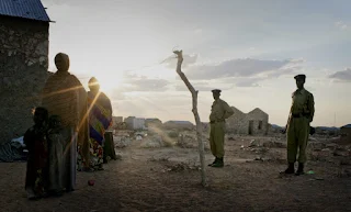 A woman and her young children stand in the evening light at an Internally Displaced People settlement in South Galkayo, Somalia with armed police standing guard.