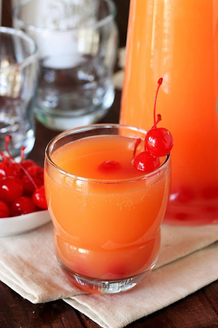 This photo shows a glass filled with Grandma's Christmas Punch, with a pitcher of the punch behind the glass. 