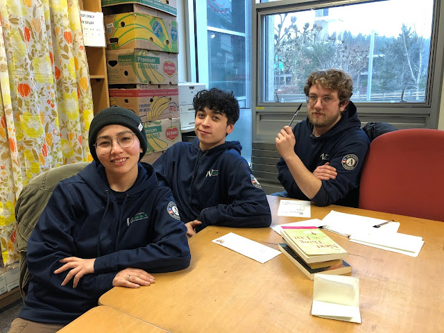 Three young adults wearing blue sweatshirts sit at an indoor table with envelopes and books on the table.