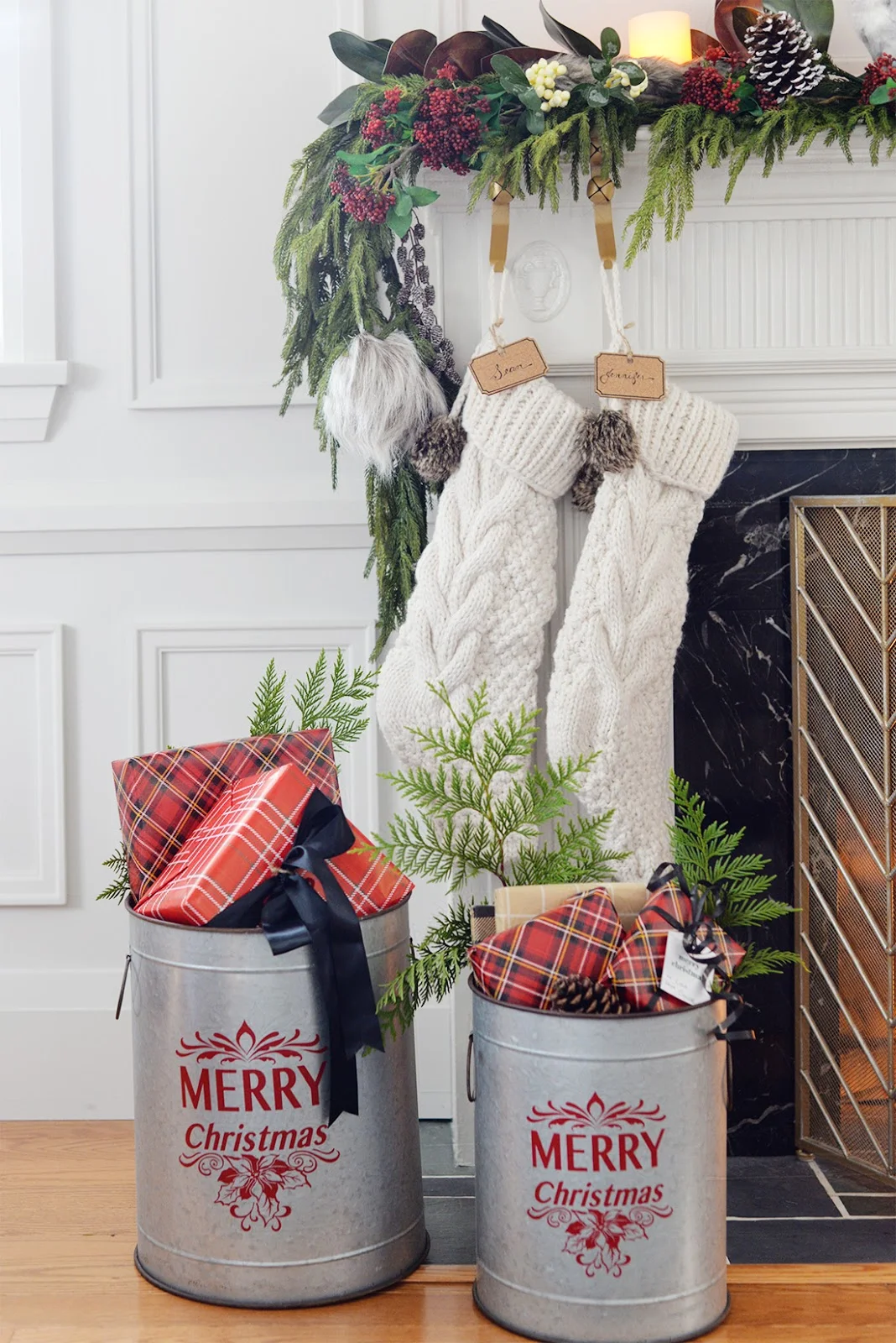 a faux christmas fireplace mantel garland with red seeded eucalyptus, a cedar garland and magnolia leaves. A galvanized bucket holding christmas gifts. Cable knit Christmas stocking