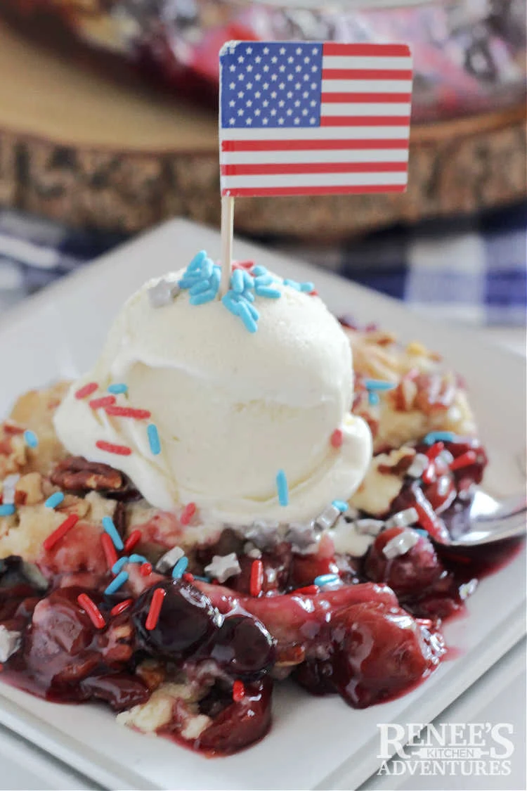 Red, White, and Blueberry Dump Cake Recipe slice on white plate with scoop of vanilla ice cream and patriotic sprinkles.