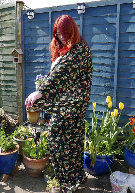 A red-haired woman in a flowery jacket and trousers. She is half-turned from the camera, examining a sleeve.