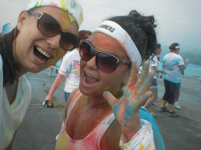 two women smiling for a photo wearing sunglasses and covered in colored powder at a color run. It's recommended you wear sunglasses to color runs to help protect your eyes.