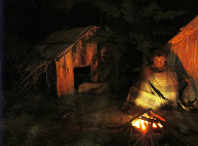 Full-scale model of a 19th century maori village, with a man sitting outside next to a fire, wrapped in a blanket.