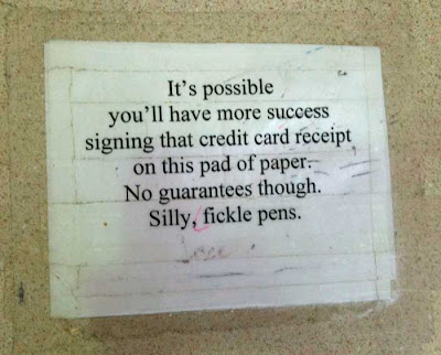Piece of paper taped to a counter. Message in Times Roman says: It's possible you'll have more success signing that credit card receipt on this pad of paper. No guarantees, though. Silly, fickle pens.