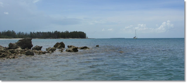 Sailboat anchored in the distance. The foreground contains a rocky sandbar and heavily treed shorelien.