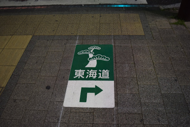 A sign on the pavement with a picture of a pine tree and an arrow pointing right. Text reads "Tokaido". In Shizuoka city
