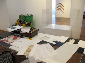 Table in a gallery in front of an empty display case. On the table are various cardboard walls and floors of miniature scenes.