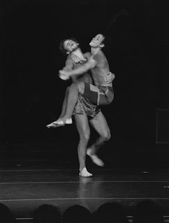 A black and white photograph of a barefoot, dancing man, holding another on a stage.