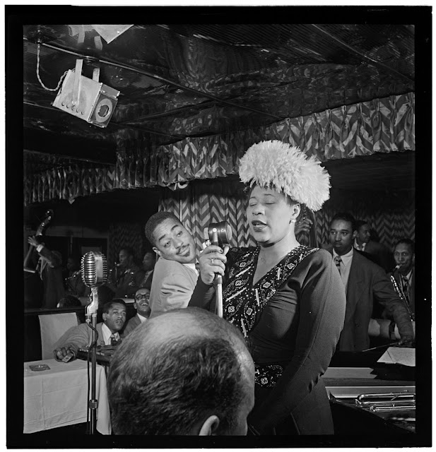 Portrait of Ella Fitzgerald, Dizzy Gillespie, Ray Brown, Milt (Milton) Jackson, and Timmie Rosenkrantz, Downbeat, New York, N.Y., ca. Sept. 1947. Caption from Down Beat: An impressive photo of a truly impressive singer Ella Fitzgerald at the Downbeat, with Dizzy Gillespie making like a faun in the background. Dizzy has gone on his own way, while Ella is still keeping the club on the beat. Forms part of: William P. Gottlieb Collection (Library of Congress). In: "Ella keeps it on the beat," Down Beat, v. 14, no. 20 (Sept. 24, 1947), p. 5.