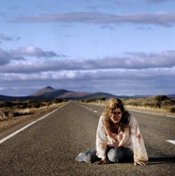 Uma moça loira sentada com as pernas dobradas no meio de uma estrada deserta. As roupas dela estão sujas de sangue e ela se apoia com as mãos no asfalto, parecendo exausta.