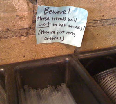 Tray of utensils, including straws, with small hand-written sign above that reads Beware! These straws will melt in hot drinks! (They're just corn, afterall)
