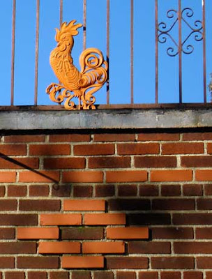 Terra cotta colored metal rooster, part of a fence above decorative brick