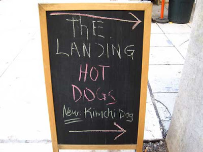 Sidewalk chalkboard sign reading The Landing Hot Dogs then New Kimchi Dogs 