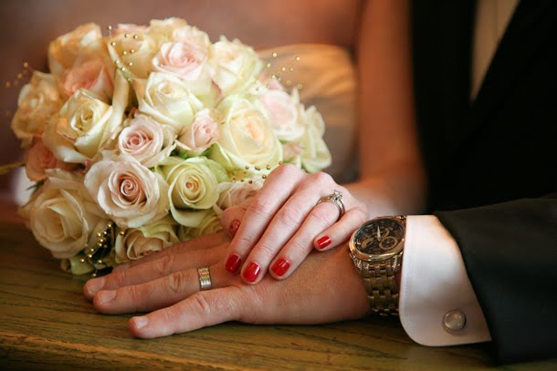 Bride and groom hold hands so you can see wedding rings with bouquet in background