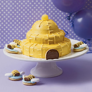 Beehive cake on a cake stand with cookies featuring bees on top.