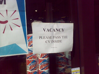 A printed sign in a shop window reading 'VACANCY  PLEASE PASS THE CV INSIDE  THANK YOU'