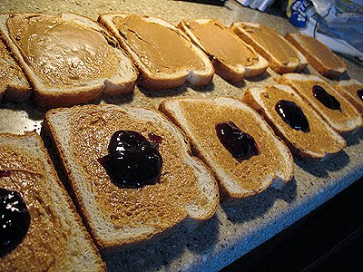 A close up photo of peanut butter spread on slices of bread and a spoonful of jelly on half of the slices.
