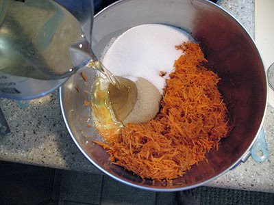 A photo of oil being added to a bowl of finely shredded carrot, eggs, granulated sugar and brown sugar.