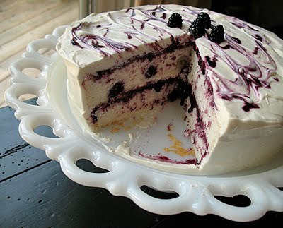 A close up photo of a vanilla bean mulberry cake on a white cake stand topped with fresh mulberries and a slice missing.