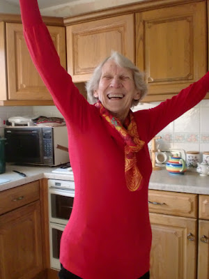 And elderly woman in a red top stands in a kitchen, a triumphant pose with arms raised and a joyful smile